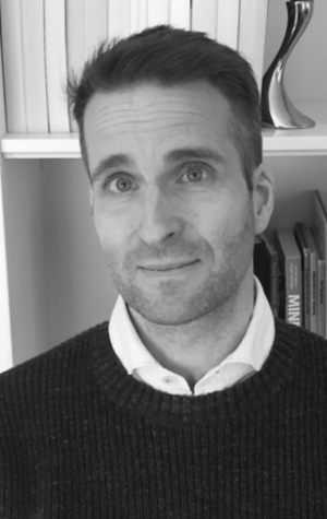 Image of Olaf Verschuren. The photo is black and white and he is standing in front of a book shelf. Olaf is wearing a dark coloured sweater.
